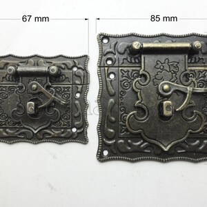 Może przedstawiać: Dwa antyczne metalowe zatrzaski w kolorze brązu. Mniejszy zatrzask ma 37 mm wysokości i 67 mm szerokości. Większy zatrzask ma 60 mm wysokości i 85 mm szerokości. Oba mają ozdobne kwiatowe wzory i zamknięcie dźwigniowe.