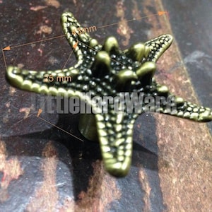 Puede incluir: Un tirador de mueble con forma de estrella de mar en metal color bronce con una superficie texturizada y rugosa. El tirador muestra líneas de medición que indican un ancho de 45 mm y una altura de 25 mm. El texto LittleHardWare es visible.