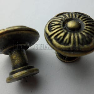 May include: Two round metal cabinet knobs with an antique brass finish. Each knob features a raised, circular, sunburst-like pattern on the face and a cylindrical base. The text LITTLEHARDWARE is visible across the center of the image.