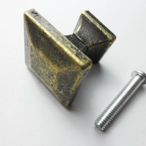 Puede incluir: Un tirador de mueble cuadrado con acabado en latón antiguo desgastado, que presenta una parte superior en forma de pirámide y una base cónica. El tirador se acompaña de un tornillo de metal plateado con cabeza plana y vástago roscado.