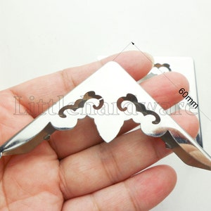 Puede incluir: Protectores de esquina de metal plateado brillante con diseños decorativos recortados. Cada esquina mide aproximadamente 60 mm. Probablemente se utilizan para proteger las esquinas de libros, cajas u otros artículos.