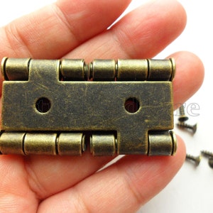 Può includere: Cerniera in ottone antico con un design decorativo. La cerniera presenta una serie di piccoli elementi decorativi rotondi lungo i bordi superiore e inferiore. La cerniera è tenuta in una mano.