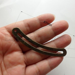 Op de afbeelding: Een gebogen metalen stuk met een gat aan het ene uiteinde. Het is 6,7 cm lang en 0,9 cm breed. Het metalen stuk is bronskleurig.