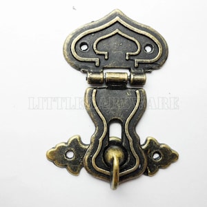 Peut inclure: Un loquet en métal bronze de style antique avec des détails ornés. Le loquet comporte une pièce supérieure décorative, un centre articulé et une section inférieure avec une petite boucle. La quincaillerie est conçue pour une armoire ou une boîte.