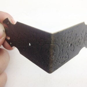 May include: A decorative corner bracket made of metal with an ornate design. The bracket is a dark bronze colour and has two holes for attaching it to a surface.