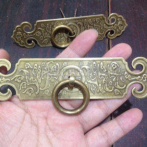 Könnte beinhalten: Zwei antike Messing-Schubladengriffe mit verzierten Designs und Ringgriffen. Die Griffe sind rechteckig mit komplizierten Mustern und chinesischen Schriftzeichen.