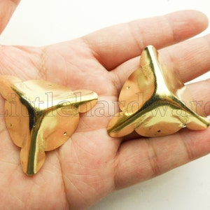 May include: Two gold-coloured metal corner protectors with rounded edges and three holes each. The protectors are designed to reinforce and decorate furniture corners.