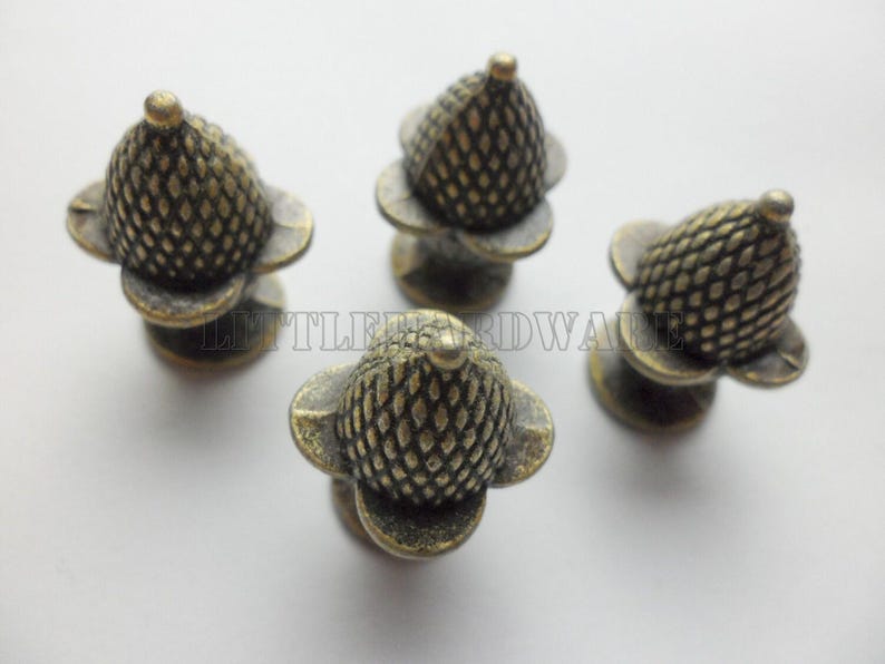 May include: Four antique brass-toned metal knobs shaped like acorns with a textured, cross-hatched pattern on the cap and a smooth base. The text LITTLEHARDWARE is visible in the center of the arrangement of these four individual hardware pieces.