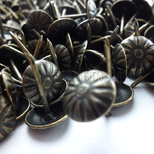 May include: A close-up of a pile of antique brass upholstery tacks with decorative heads. The tacks have a starburst design and are perfect for adding a touch of vintage charm to furniture, crafts, and home decor.