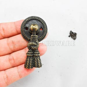 Puede incluir: Un tirador de cajón circular de latón antiguo con un borde de textura de cuerda y un asa central en forma de borla decorativa con un nudo. El herraje tiene un acabado oscuro y desgastado e incluye dos pequeños tornillos de montaje.