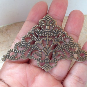 May include: A silver-toned metal corner embellishment with a decorative, intricate, and ornate design. The embellishment is shaped like a triangle with a pointed top and rounded edges.