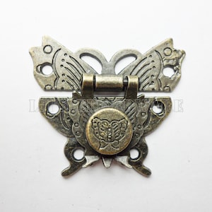 May include: A bronze butterfly-shaped latch with a decorative design. The latch has a hinged section and a round, decorative knob in the center.