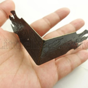 May include: A dark brown metal corner bracket with an ornate design. The bracket is bent at a right angle and has decorative cutouts. The text "Littlehardware" is visible in the image. Ideal for furniture or craft projects.