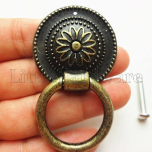 May include: A circular bronze-toned metal drawer pull featuring a central flower motif with a beaded border and a swinging ring handle. The hardware includes a separate silver-coloured mounting screw for installation on furniture or cabinetry.