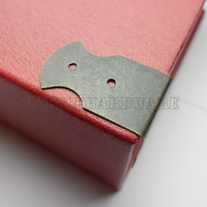 Puede incluir: Primer plano de un libro encuadernado en cuero rojo con una pieza de esquina decorativa de latón. La pieza de esquina tiene dos agujeros y un diseño único.