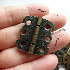 Puede incluir: Una bisagra de color bronce antiguo, de aproximadamente 27 mm de ancho y 29 mm de largo, con detalles decorativos. La bisagra tiene un pasador central y múltiples orificios para tornillos para la fijación. La imagen muestra la bisagra en una mano.