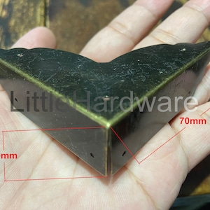Puede incluir: Una escuadra de esquina de metal color bronce, de 70 mm por 20 mm, con un diseño decorativo. El soporte tiene un acabado envejecido y se muestra en una mano. El texto "Little Hardware" es visible.