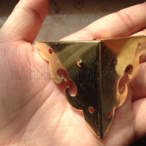 May include: Gold metal corner bracket with a decorative design. The bracket has a triangular shape with rounded edges and a floral pattern.