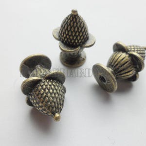 May include: Three antique brass-toned metal knobs shaped like acorns with a textured, cross-hatched cap and a smooth, fluted base. Each knob features a small, circular mounting point at the bottom. The word HARDWARE is visible in faint text.