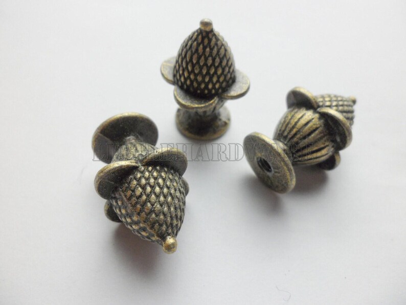 May include: Three antique brass-toned metal knobs shaped like acorns with a textured, cross-hatched cap and a smooth, fluted base. Each knob features a small, circular mounting point at the bottom. The word HARDWARE is visible in faint text.