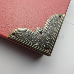 Puede incluir: Primer plano de una pieza de esquina decorativa para un libro o una caja. La pieza de esquina está hecha de metal y tiene un acabado marrón oscuro. Presenta un intrincado diseño floral.