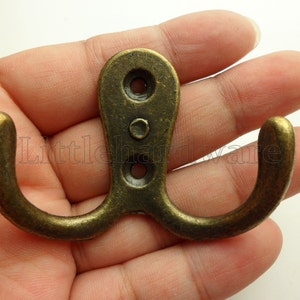 May include: Antique brass double hook with a curved design. The hook has a rectangular base with two screw holes and a central decorative rivet. The curved hooks extend outwards, offering a vintage aesthetic.