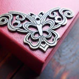 Può includere: Un elemento decorativo in metallo a forma di farfalla color bronzo antico. La farfalla presenta dettagli traforati e complessi ed è posta su una superficie rossa. Il design presenta ali simmetriche con motivi a spirale.