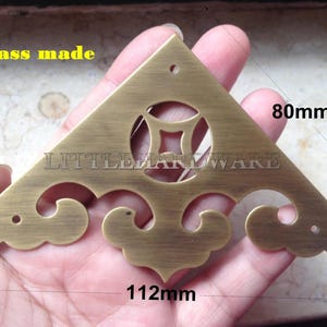 Könnte beinhalten: Eckwinkel aus Messing mit dekorativen Schnörkeln und einem mittigen Ausschnitt. Die Halterung ist 11,2 cm lang und 8 cm breit. Der Text "brass made" ist sichtbar. Das Produkt ist von "LITTLE HARDWARE".