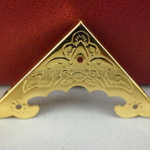 May include: Gold metal corner protector with an ornate floral design. The protector has two holes on each side.