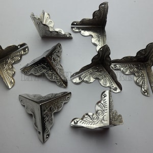 May include: Eight silver corner protectors with a decorative floral design. The corner protectors are triangular in shape and have a smooth, shiny finish.