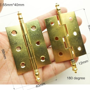 Può includere: Due cerniere in metallo dorato con terminali decorativi. Ogni cerniera misura 4,5 cm per 4 cm e ha un perno di 1,2 cm di diametro. Le cerniere sono progettate per aprirsi di 180 gradi.