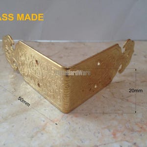 May include: A brass corner bracket with decorative floral patterns. The bracket measures 50mm in length and 20mm in width. The text "BRASS MADE" is visible. The bracket has pre-drilled holes for fixing.