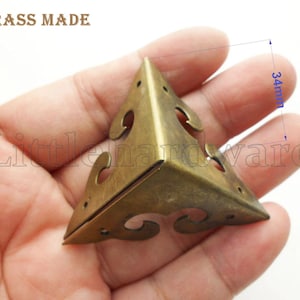 May include: A brass corner bracket, 3.4cm in length, held in a hand. The bracket is triangular with decorative cutouts and small holes. The text "BRASS MADE" is visible at the top.