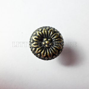 May include: A round, antique brass-colored cabinet knob with a floral design. The knob has a detailed, textured surface and is centered on a white background. The text "LITTLE HARDWARE" is visible.