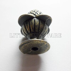 May include: A bronze-toned metal cabinet knob featuring a textured, pineapple-shaped top with a fluted base and a circular mounting plate. The text LITTLE HARDWARE is visible across the center of the object in a light, outlined font.