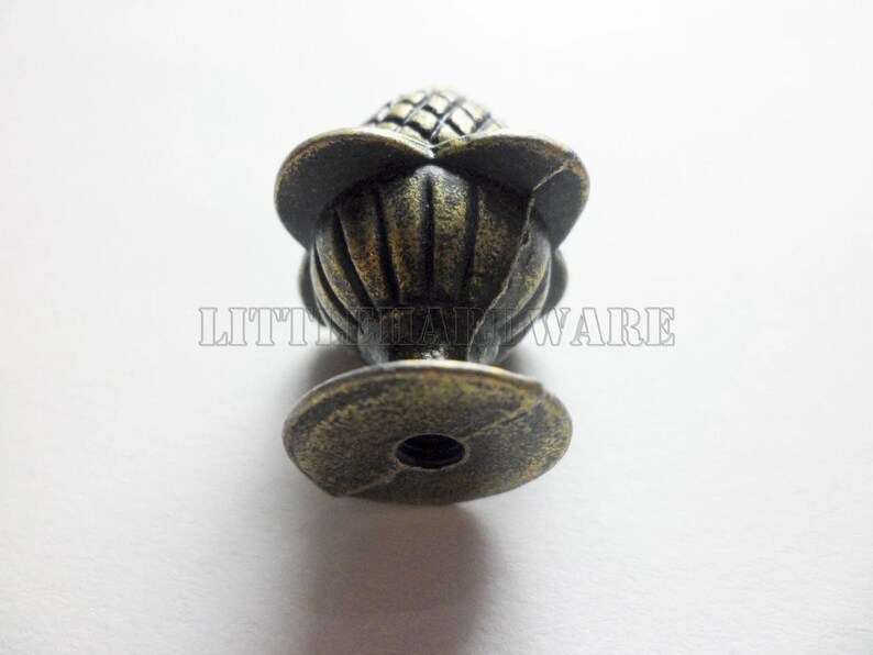 May include: A bronze-toned metal cabinet knob featuring a textured, pineapple-shaped top with a fluted base and a circular mounting plate. The text LITTLE HARDWARE is visible across the center of the object in a light, outlined font.