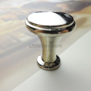May include: A polished silver-tone metal cabinet knob with a smooth, rounded top and a tapered, flared base. The hardware features a reflective, high-gloss finish and the text Littlehardware is visible on the central stem of the object.