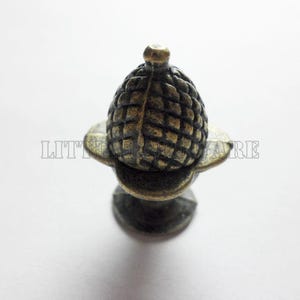 May include: A bronze-toned metal cabinet knob shaped like an acorn with a textured, cross-hatched cap and a smooth base. The hardware features a dark, weathered finish with highlights on the raised edges, providing a distinct, aged appearance.