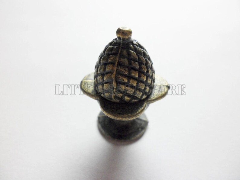 May include: A bronze-toned metal cabinet knob shaped like an acorn with a textured, cross-hatched cap and a smooth base. The hardware features a dark, weathered finish with highlights on the raised edges, providing a distinct, aged appearance.