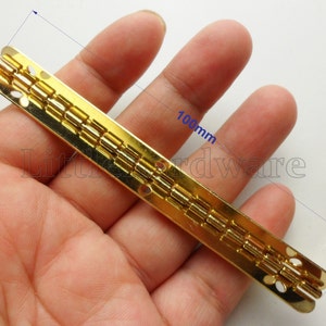 Può includere: Una cerniera rettangolare dorata, lunga circa 10 cm e larga 9 mm, è tenuta in mano. La cerniera è composta da più segmenti collegati. Piccole viti dorate sono visibili sullo sfondo. Immagine di un componente hardware.