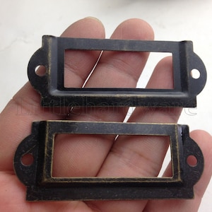 Puede incluir: Dos portaetiquetas de cajón de latón antiguo con una ventana rectangular para insertar una etiqueta. Los soportes tienen una parte superior e inferior redondeadas y un agujero a cada lado.