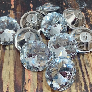 May include: A collection of clear, faceted crystal buttons with silver metal backs. The buttons are round and have a shiny, reflective surface. The image is taken on a wooden surface.