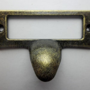 Puede incluir: Portaetiquetas de latón antiguo con marco rectangular y pomo redondeado. El portaetiquetas tiene dos pequeños orificios para el montaje. El metal tiene un acabado envejecido, lo que le da un aspecto vintage.