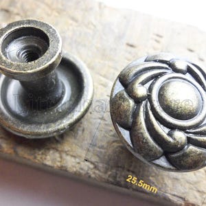 Puede incluir: Dos tiradores de armario de bronce antiguo. Uno mide 22 mm de alto, el otro 25,5 mm de ancho, con un diseño floral ornamentado. Los tiradores tienen un acabado envejecido, lo que sugiere una apariencia antigua. El herraje es probablemente para muebles.