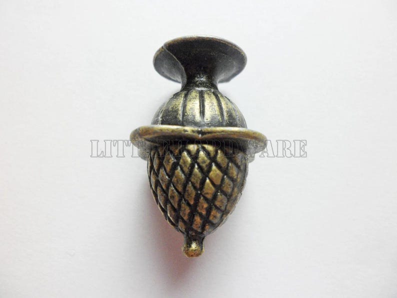 May include: A metallic acorn-shaped cabinet knob with an antique brass finish. The knob features a textured, cross-hatched pattern on the lower section and a smooth, rounded cap. The text LITTLE HARDWARE is visible across the center of the object.