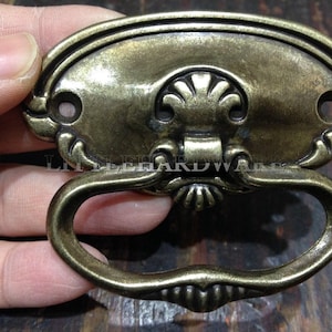 Puede incluir: Un tirador de cajón de metal en tono latón antiguo con una placa ovalada decorada con motivos de concha y dos orificios para tornillos. Un asa abatible ovalada y articulada cuelga del centro. El texto LITTLEHARDWARE está grabado en el centro.