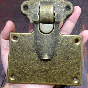 Puede incluir: Cierre metálico de color latón antiguo con acabado texturizado. El cierre presenta una base rectangular y una pieza superior decorativa. El mecanismo central incluye una barra articulada y un pestillo curvado. Ideal para un baúl o cofre.