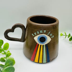 May include: A brown ceramic mug with a heart-shaped handle. The mug features an eye design with rainbow rays and the words "trans joy". The mug is a unique and artistic piece.