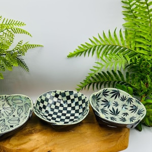 May include: Three small ceramic bowls with different designs. The bowls are on a wooden tray. The designs are geometric shapes, a checkerboard pattern, and a leaf pattern.