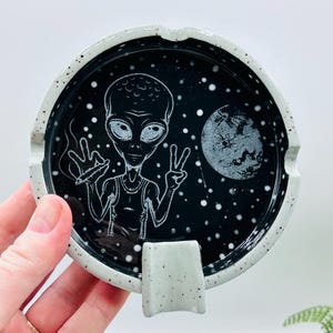 May include: A black ceramic ashtray with a white speckled rim and a built-in rest. The ashtray features a white illustration of an alien with a peace sign and a lit cigarette, with a moon and stars in the background.